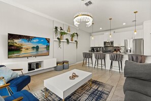 A modern living room with a flat-screen TV, a white coffee table, and a blue sofa. There is a kitchen area with white cabinets and stainless steel appliances. - Mickey & Guardians Theme • Splash Pool • Waterpark • Golf • Resort • Near Disney (Davenport)