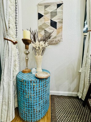 Una habitación de esquina con una mesa redonda de mosaico azul, una vela y un florero con plantas secas. Un tapiz de diseño geométrico cuelga en la pared.