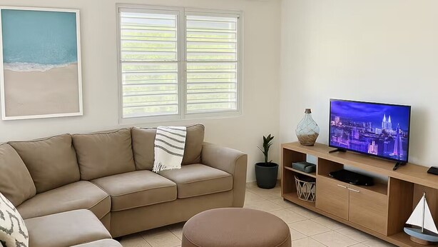 A living room with a beige sofa, a round ottoman, a TV on a wooden stand, and a cityscape painting on the wall. - Minutes to EVERYTHING! Gated home! AC in all rooms! (Hatillo)