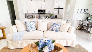 A living room with a beige sofa, a round wooden coffee table with a woven tray and blue flowers, and a kitchen in the background. - Beautiful Cape Coral Getaway with Sparkling Pool & Relaxing Outdoor Space! (Cape Coral)