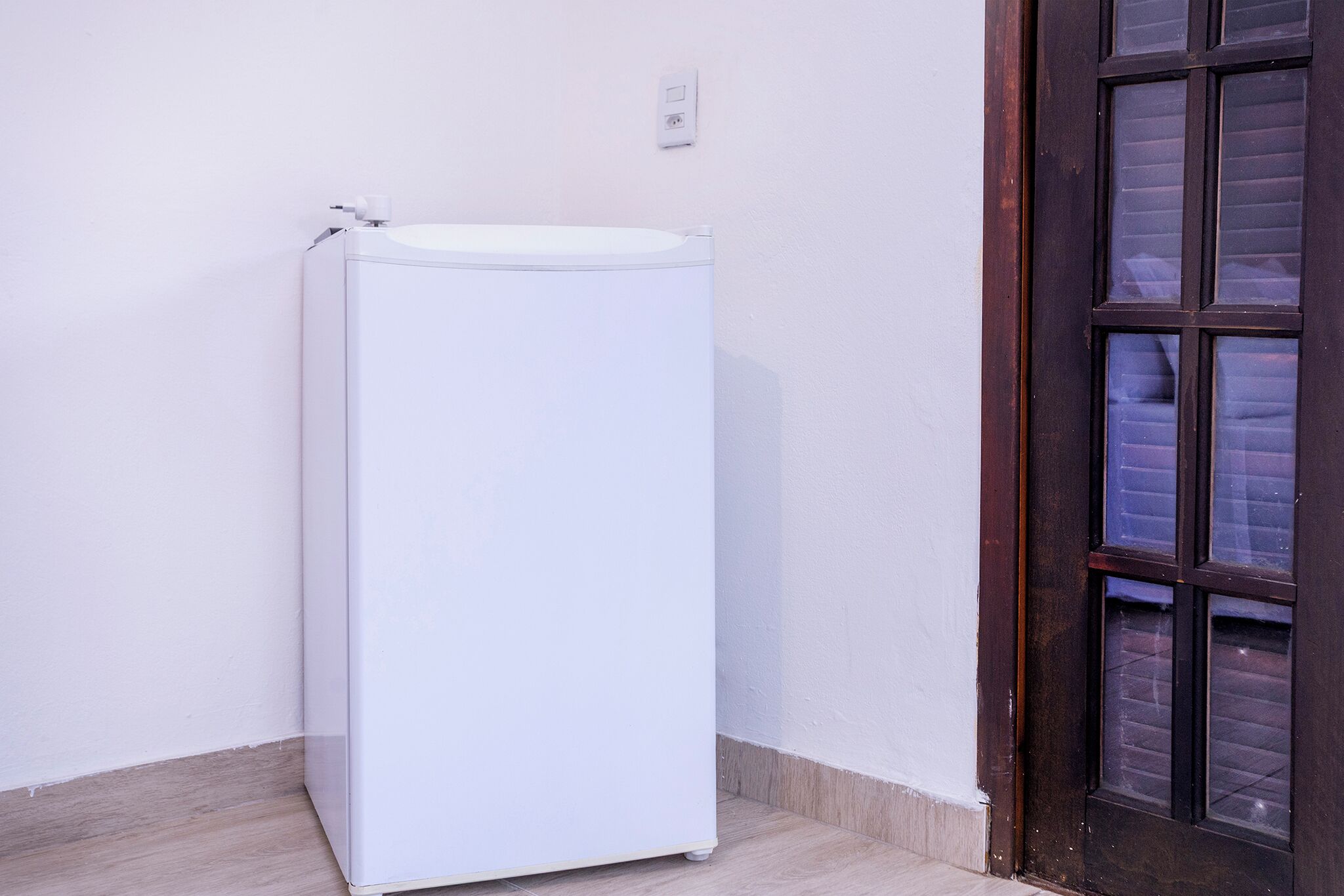 Un refrigerador blanco en un rincón de una habitación con piso de madera y una puerta de madera oscura con paneles de vidrio.