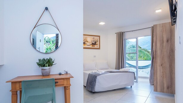 A bedroom with a bed, a desk with a chair, a potted plant, a hanging mirror, and a painting. - Pousada Cahê (Ilhabela)