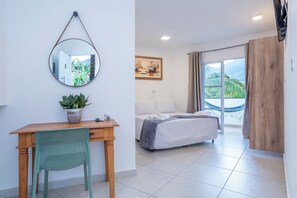 A bedroom with a bed, a desk with a chair, a potted plant, a hanging mirror, and a painting. - Pousada Cahê (Ilhabela)