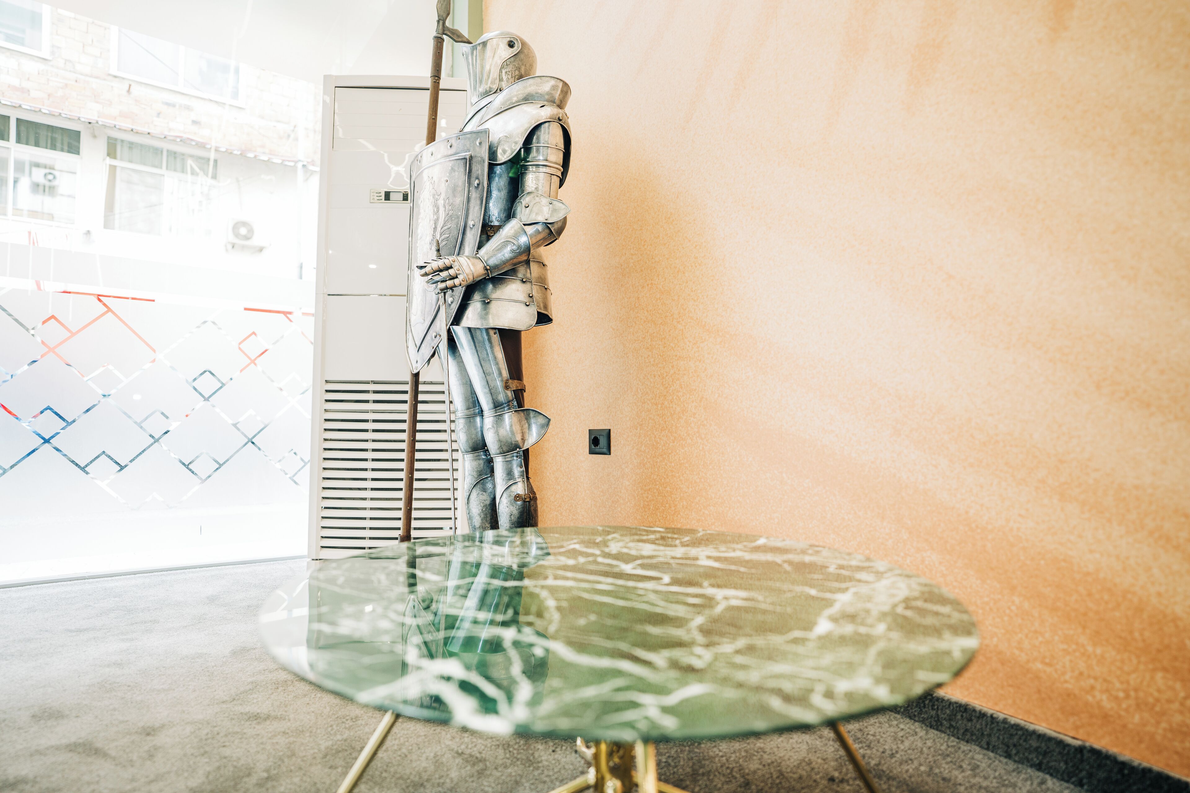 A room with a glass table, a mannequin in armor, and a wall with a geometric pattern.