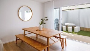 A modern dining area with a wooden table and benches, a round mirror on the wall, and a view of a backyard with a white fence and outdoor seating.