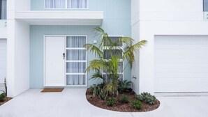 A modern two-story house with a light blue exterior, a front door with a glass panel, and a small garden with palm trees and shrubs. - Olas Villa 2 | Luxe 3BR · Plunge Pool · Near Beach (Anna Bay)