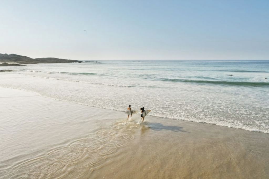 A beach scene with two people walking along the shore, carrying surfboards.