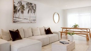 A modern living room with a sectional sofa, a wooden coffee table, and a painting of palm trees. - Olas Villa 4 | Luxe 3BR · Plunge Pool · Near Beach (Anna Bay)