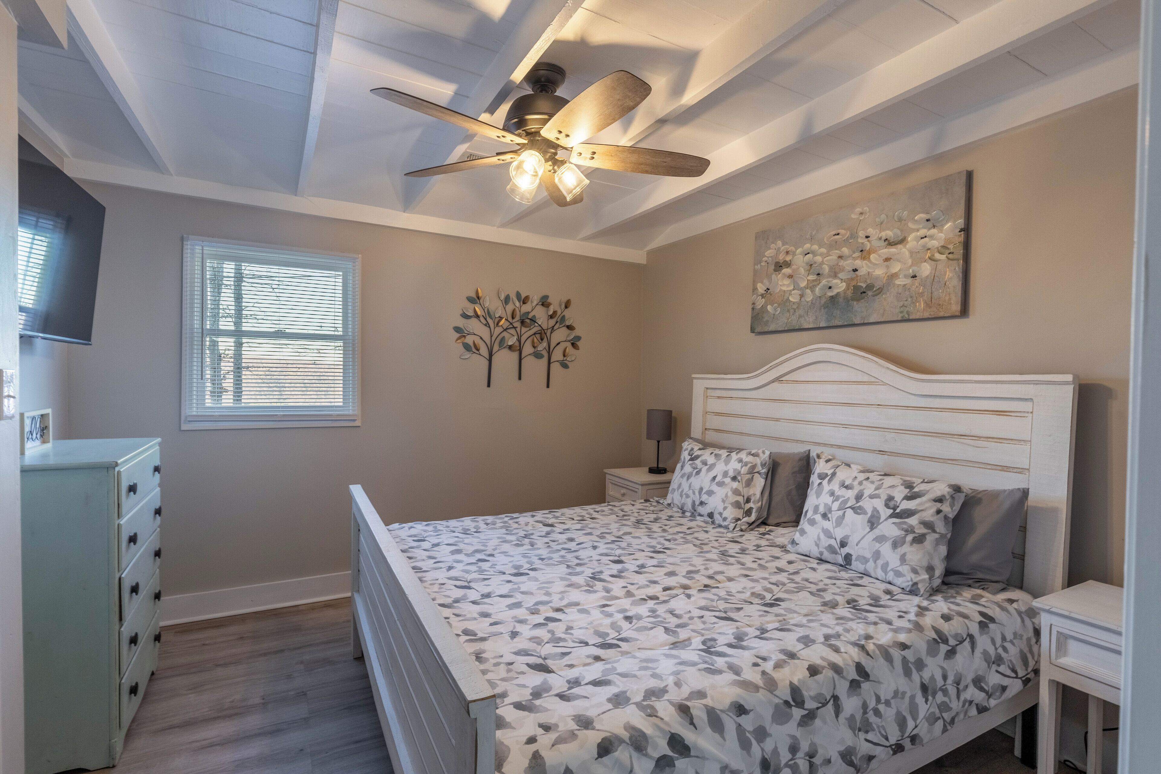 Ein Schlafzimmer mit einem Bett, einem Deckenventilator, einem Fenster mit Jalousien, einem Schminktisch und einem Wandbild.