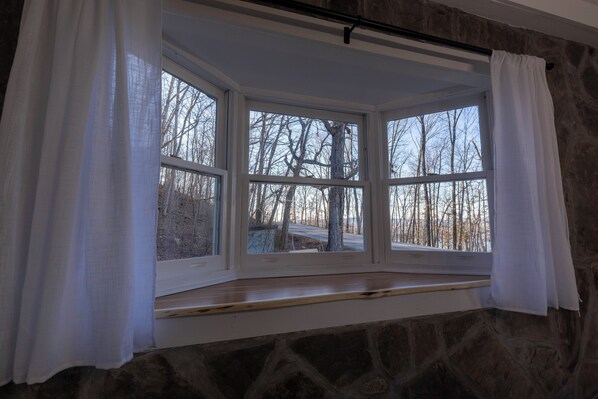 A bay window with white curtains, stone walls, and a view of leafless trees.