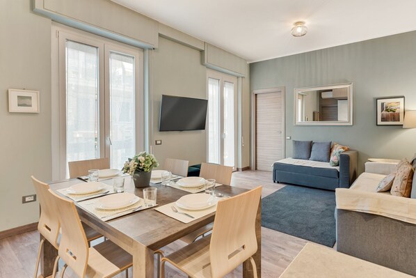 Un salon moderne comprenant un coin repas, un canapé, un téléviseur fixé au mur et une grande fenêtre.