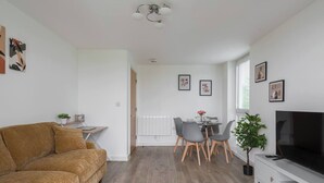 A modern living room with a brown sofa, a dining table with chairs, a TV, and a radiator. - Beautiful Flat • Prime Location • Balcony • Free Parking • Self-Check-In (Milton Keynes)