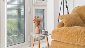 A room with a beige sofa, a small round table with a vase of flowers, and a window with blinds. - Beautiful Flat • Prime Location • Balcony • Free Parking • Self-Check-In (Milton Keynes)