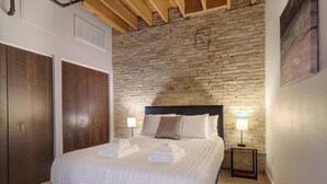 Un dormitorio con pared de piedra, una cama con ropa blanca, cabecera de madera y una mesita de noche con lámpara.