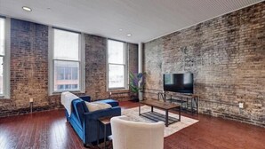 Un salón amplio con pared de ladrillo, un sofá azul, un sillón blanco y un televisor de pantalla plana.