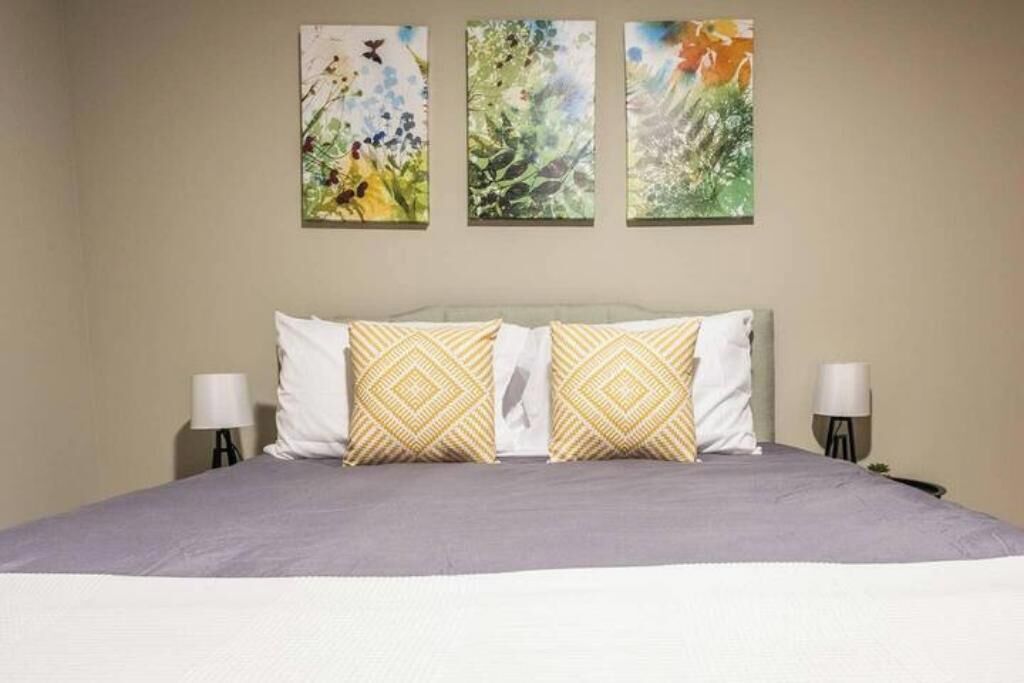 A bed with a gray bedspread, two white pillows, and two patterned pillows. There are three wall-mounted paintings above the bed.