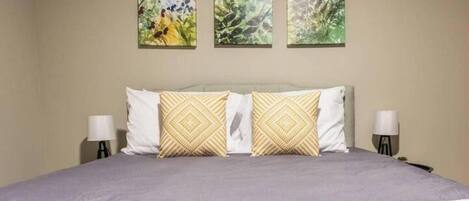 A bed with a gray bedspread, two white pillows, and two patterned pillows. There are three wall-mounted paintings above the bed.