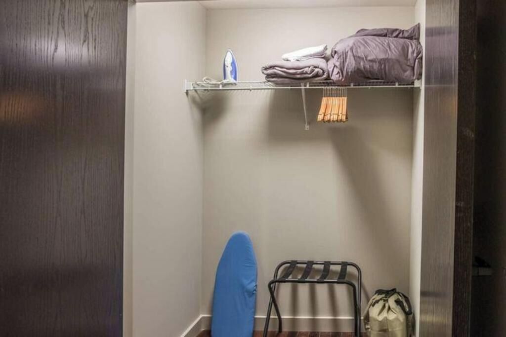 A compact room with a metal rack holding folded clothes, a blue ironing board, and a bag on the floor.