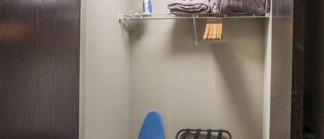 A compact room with a metal rack holding folded clothes, a blue ironing board, and a bag on the floor.