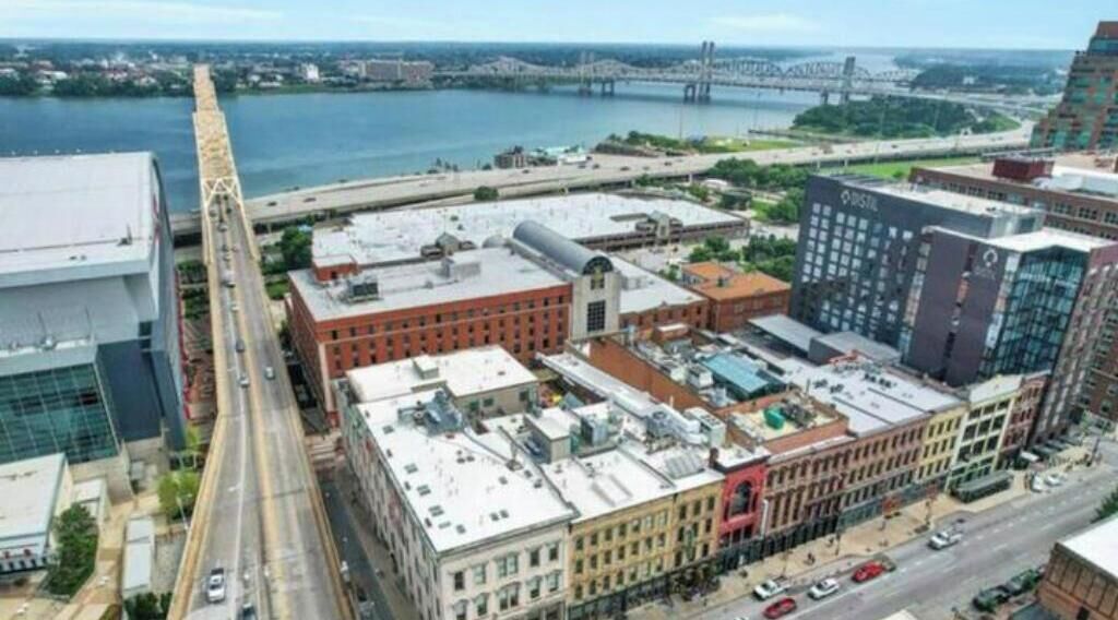 An aerial view of a cityscape with a river, a bridge, and various buildings including a large red brick building and a modern glass building.