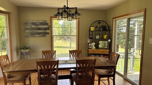 A dining area with a wooden table and chairs, a chandelier, a bookshelf with plants, and a large window with a view of greenery. - Peaceful country 5 bedroom home. Come make memories at your home away from home. (Poynette)