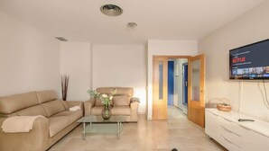 A modern living room with a beige sofa, a glass coffee table, and a flat-screen TV mounted on the wall.