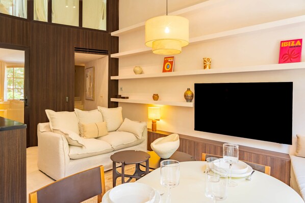 A modern living room with a sofa, a dining table with glasses, a TV mounted on the wall, and a bookshelf with decorative items.