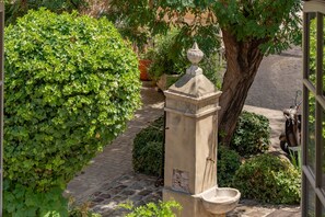 A stone fountain with a basin, surrounded by lush green foliage and a paved pathway.