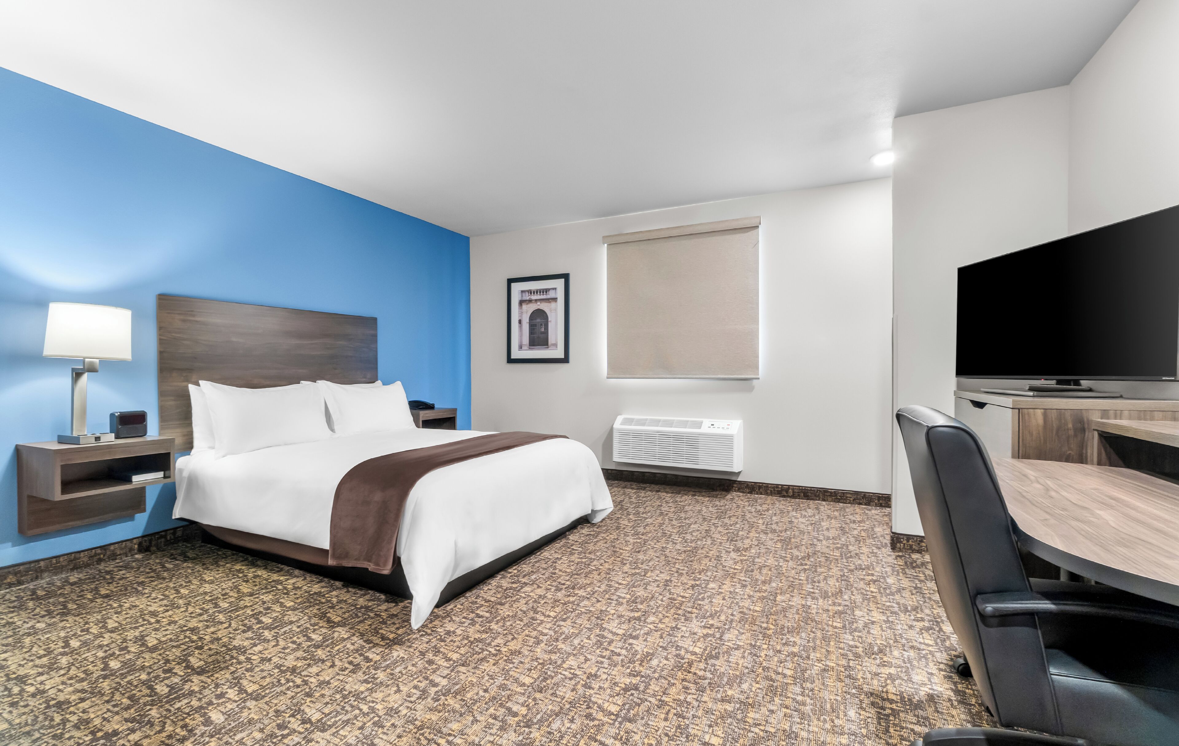 A hotel room with a bed, a desk with a chair, a television, a lamp, a framed picture, and a window with a blind.
