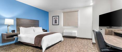 A hotel room with a bed, a desk with a chair, a television, a lamp, a framed picture, and a window with a blind.