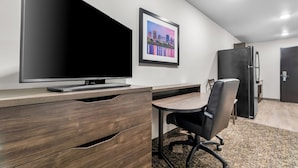Une chambre d’hôtel moderne équipée d’un téléviseur à écran plat, d’un tableau représentant un paysage urbain encadré, d’un bureau en bois, d’un fauteuil de bureau noir, d’un réfrigérateur et d’une porte.