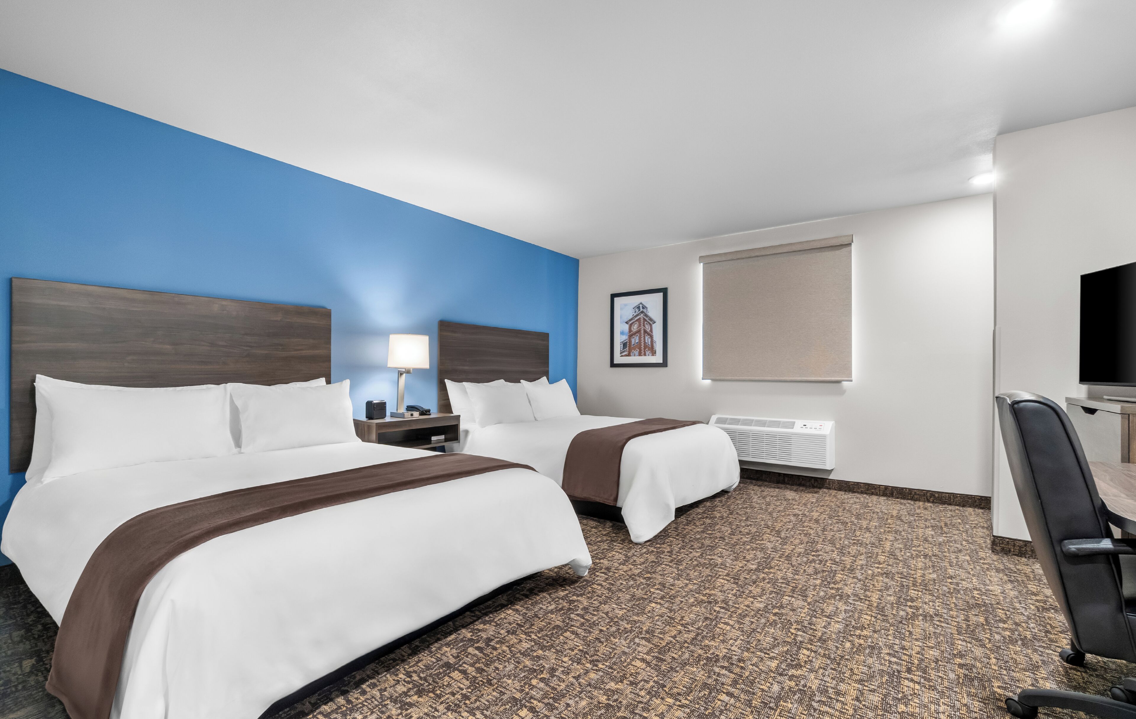 A hotel room with two beds, a desk, a chair, a lamp, and a picture on the wall.