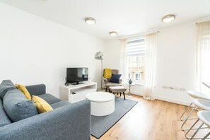 Un salon moderne avec un canapé gris, un téléviseur à écran plat, une table basse blanche et une fenêtre avec des rideaux.