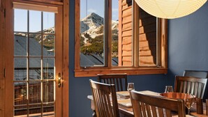 A dining area with a table set for two, a wooden chair, and a view of a mountain through a window. - Big Sky Ski Chalet 3 Mins to Lifts with Hot Tub, Fireplace & Queen Bed Bunk Room (big sky)