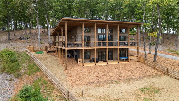 A two-story wooden cabin with a balcony, surrounded by trees and a fenced yard. - Modern Rustic Log Cabin-Dog Friendly-Fenced-Views-Hot Tub (Morganton)
