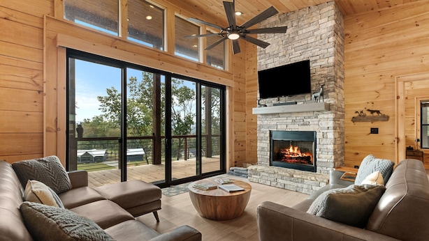 A spacious living room with a stone fireplace, leather sofas, and a large sliding glass door leading to an outdoor deck. - Modern Rustic Log Cabin-Dog Friendly-Fenced-Views-Hot Tub (Morganton)