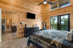 A bedroom with a large bed, a ceiling fan, a flat-screen TV, and a view of the outdoors.
