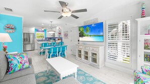 A bright living room with a TV, a ceiling fan, and a kitchen in the background.