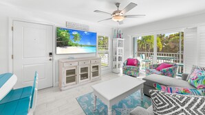 A bright living room with a white ceiling fan, a large flat-screen TV, and a view of the outdoors through large windows.