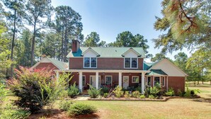 A two-story brick house with a green roof, a covered porch, and a well-maintained front yard with various plants and shrubs.