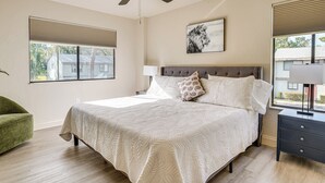 Quarto com cama, ventilador de teto, janela com persianas, mesa de cabeceira e uma cadeira verde.