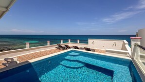 En pool på taket med kristallklart blått vatten, ett vitt kaklat däck och utsikt över havet.