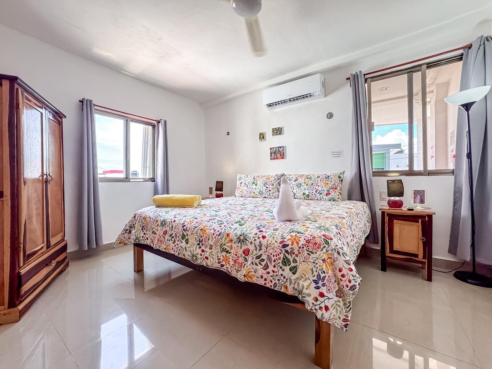 A hotel room with a bed, a wardrobe, an air conditioning unit, a bedside table, a lamp, and a window with curtains.