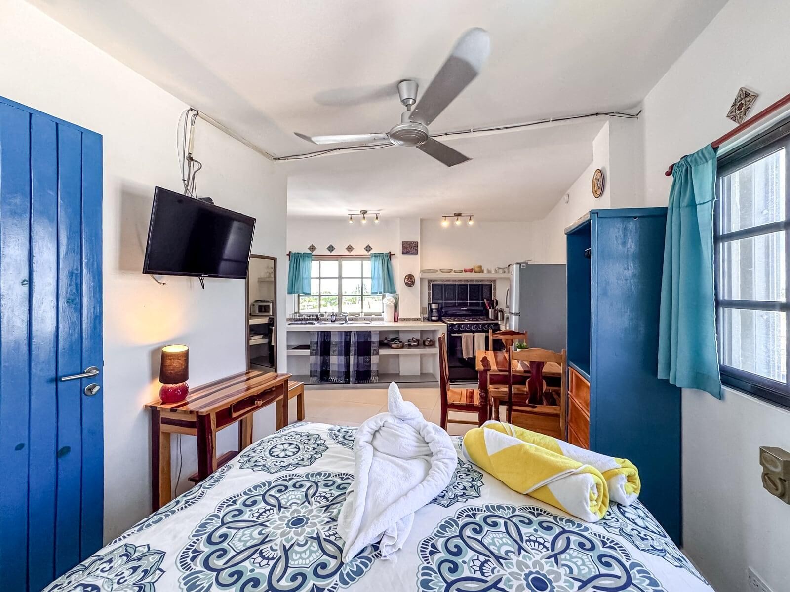 Une pièce de taille modeste comprenant un lit, un bureau, une télévision, un ventilateur de plafond et une kitchenette.