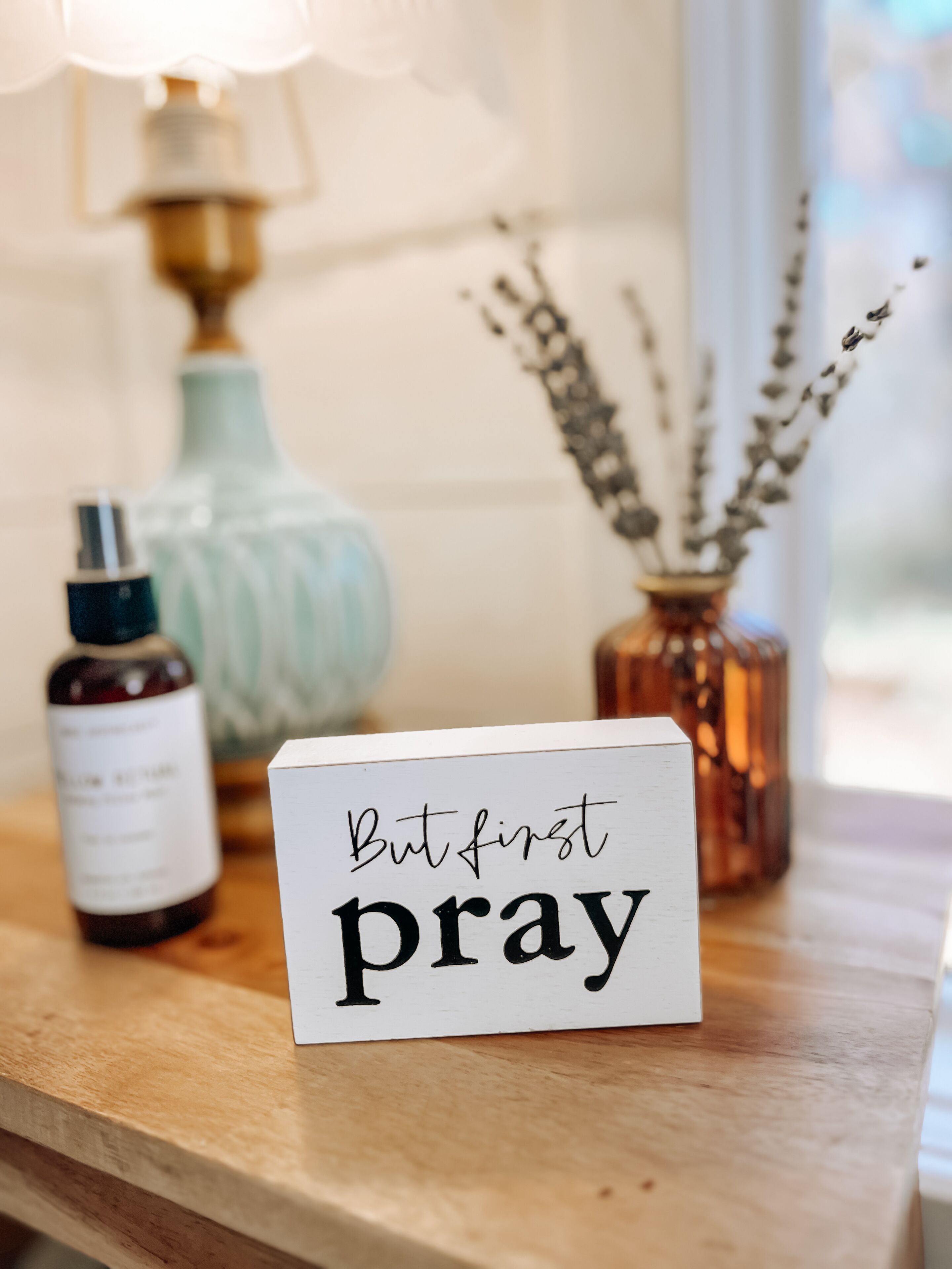 Una mesa de madera con un letrero que dice 'But first pray', una botella, un florero con flores secas y una lámpara al fondo.
