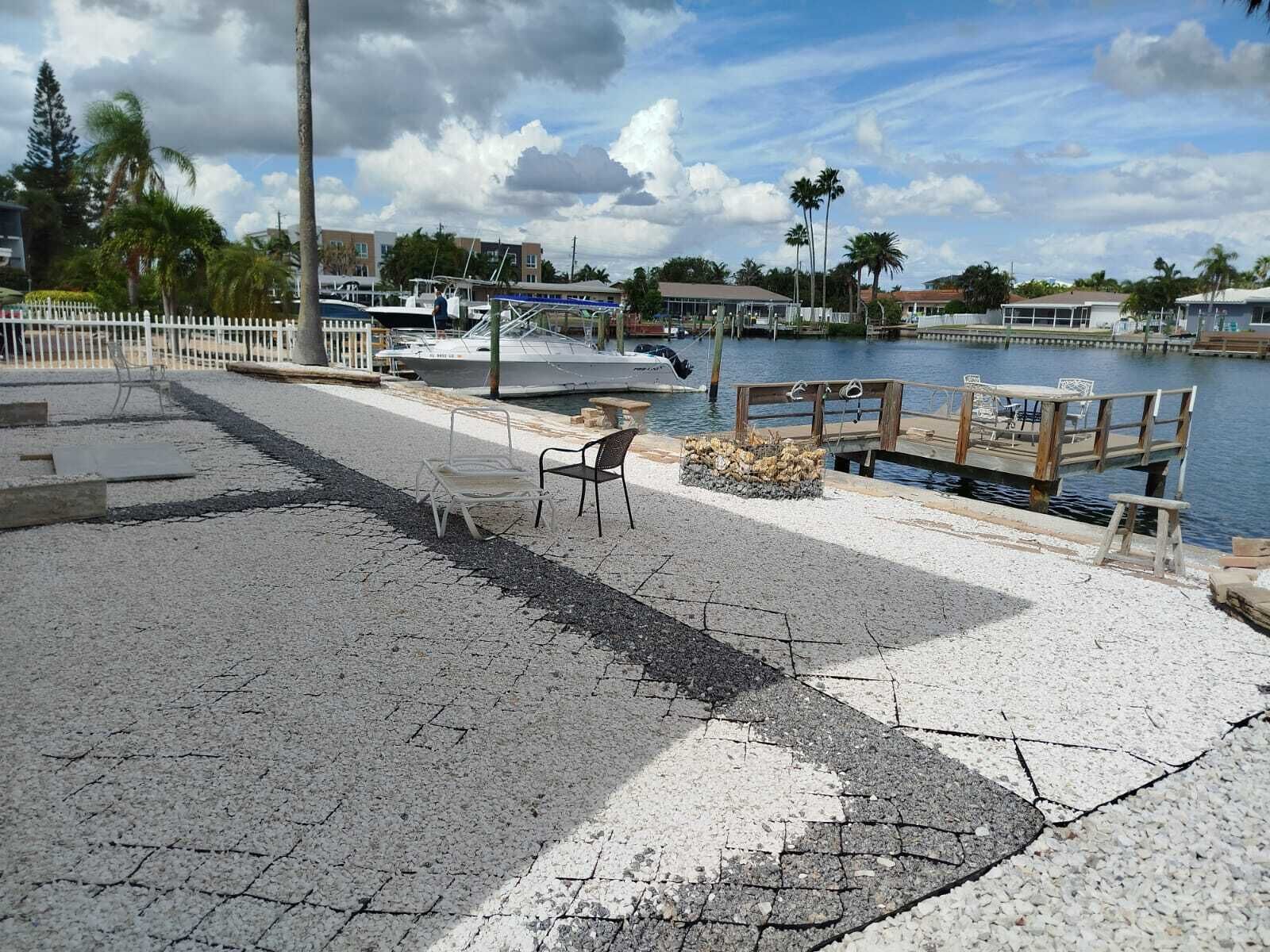 Un paseo marítimo con un banco y un pequeño hogar para fuego, con vistas a un puerto deportivo con barcos y palmeras.