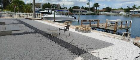 Un paseo marítimo con un banco y un pequeño hogar para fuego, con vistas a un puerto deportivo con barcos y palmeras.
