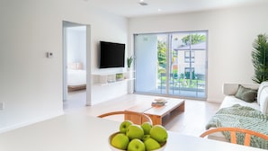 A modern living room with a flat-screen TV, a dining area with a bowl of green apples, and a large sliding glass door leading to a balcony with a view of greenery and a building. - Beach Haus Key Biscayne II (Key Biscayne)