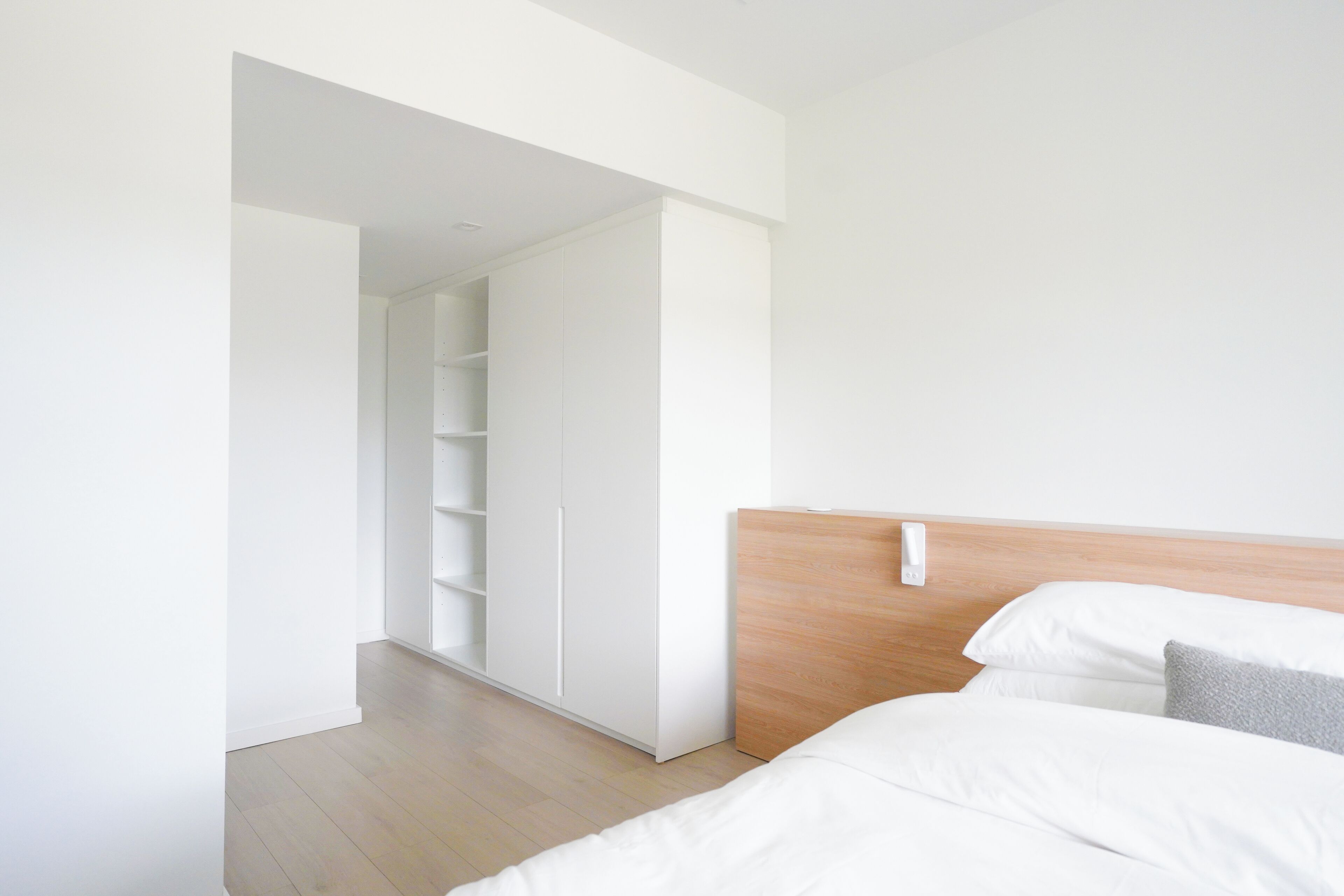 A modern bedroom with a wooden headboard, white bedding, and a built-in wardrobe.