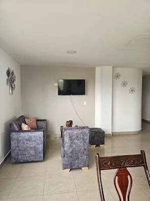 Una sala de estar con un televisor de pantalla plana montado en la pared, un sofá gris con cojines, una mesa de centro de madera y una silla de madera.